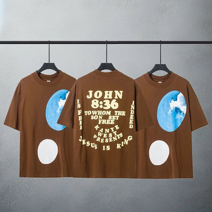 Brown t-shirt with 'John 8:36' verse and 'Jesus is King' text in a circular design, flanked by half-moon and earth graphics, representing Kanye West's spiritual and musical journey.