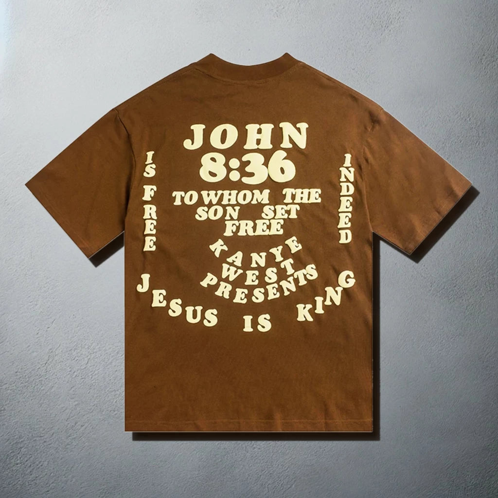 Brown t-shirt with 'John 8:36' verse and 'Jesus is King' text in a circular design, flanked by half-moon and earth graphics, representing Kanye West's spiritual and musical journey.