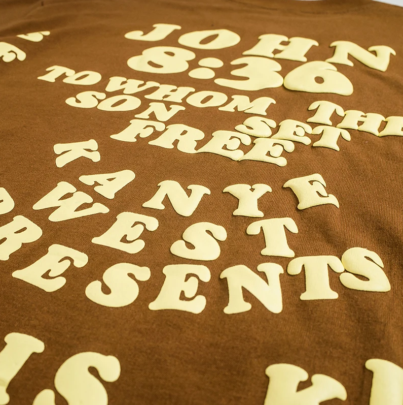 Close up Brown t-shirt with 'John 8:36' verse and 'Jesus is King' text in a circular design, flanked by half-moon and earth graphics, representing Kanye West's spiritual and musical journey.